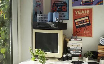 Mesa decorada com itens retrô, incluindo monitor antigo, fitas VHS e pôsteres vintage na parede, destacando o estilo de decoração retrô.