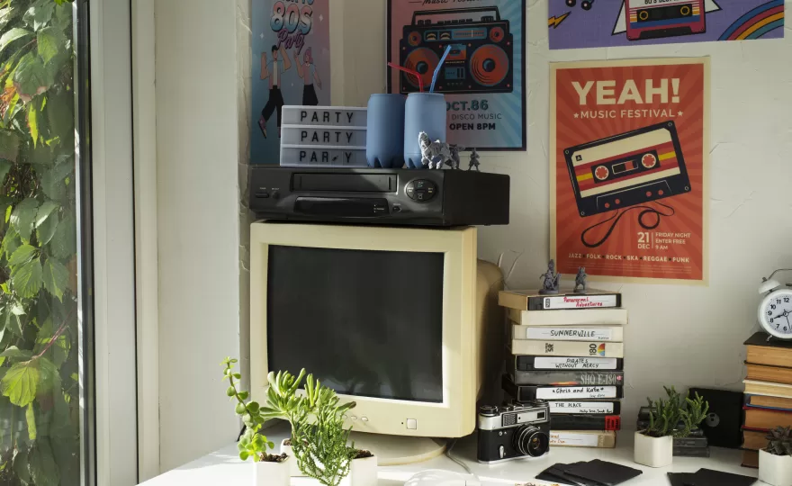 Mesa decorada com itens retrô, incluindo monitor antigo, fitas VHS e pôsteres vintage na parede, destacando o estilo de decoração retrô.